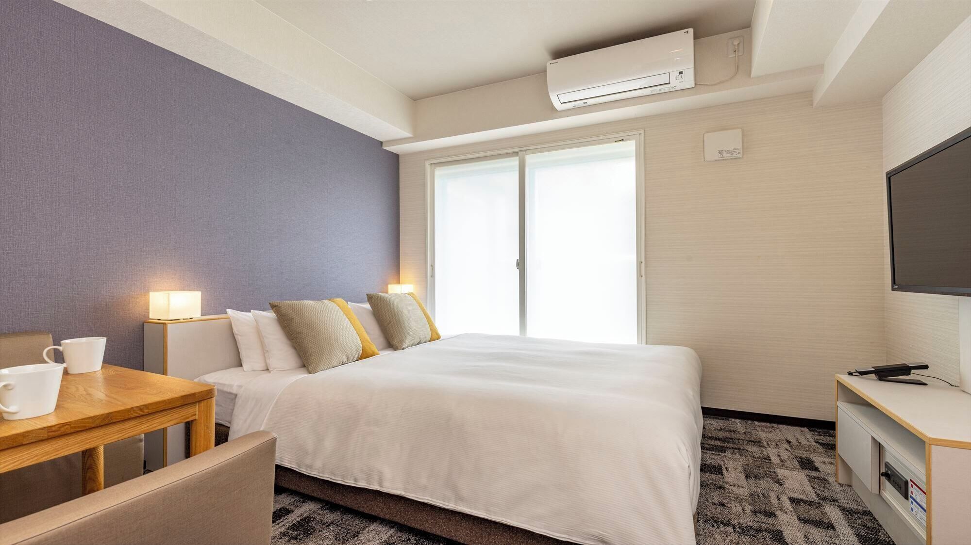 Een hotelkamer met een bed, nachtkastje, lamp, televisie en een raam.