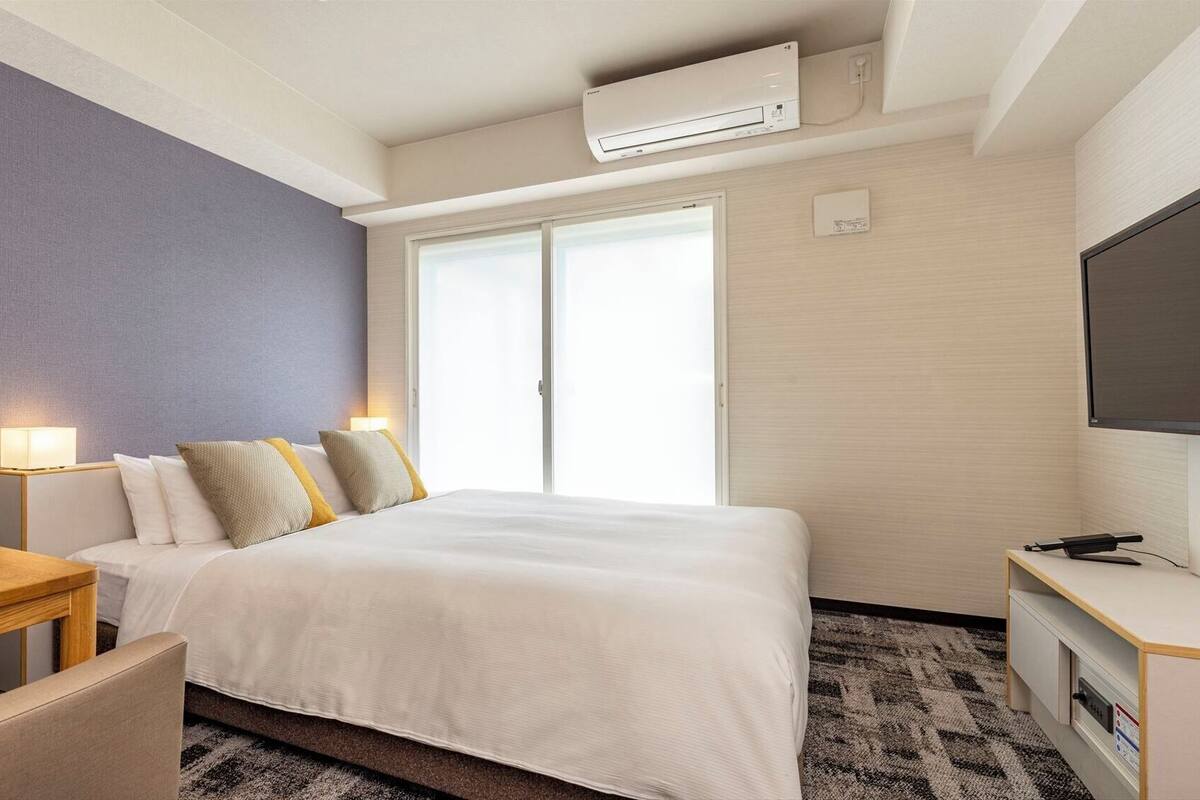 A hotel room with a bed, bedside table, lamp, TV, and a window.