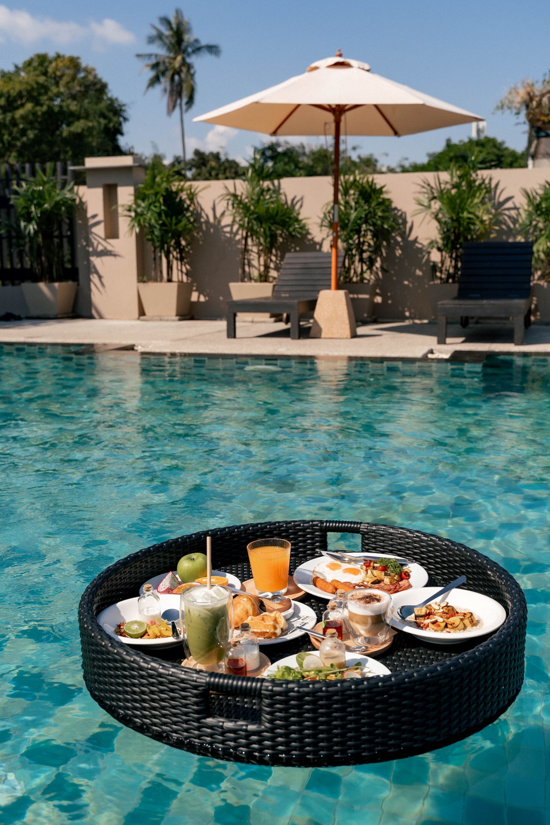 A poolside dining setup with a wicker tray holding plates of food and drinks, an umbrella, and lounge chairs in the background.