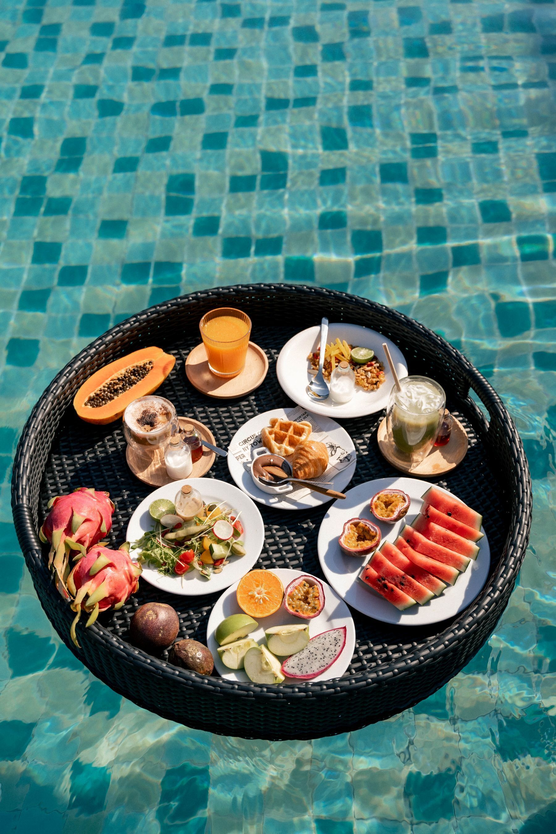 A round wicker tray with various breakfast items including fruits, waffles, and beverages, placed on a poolside.