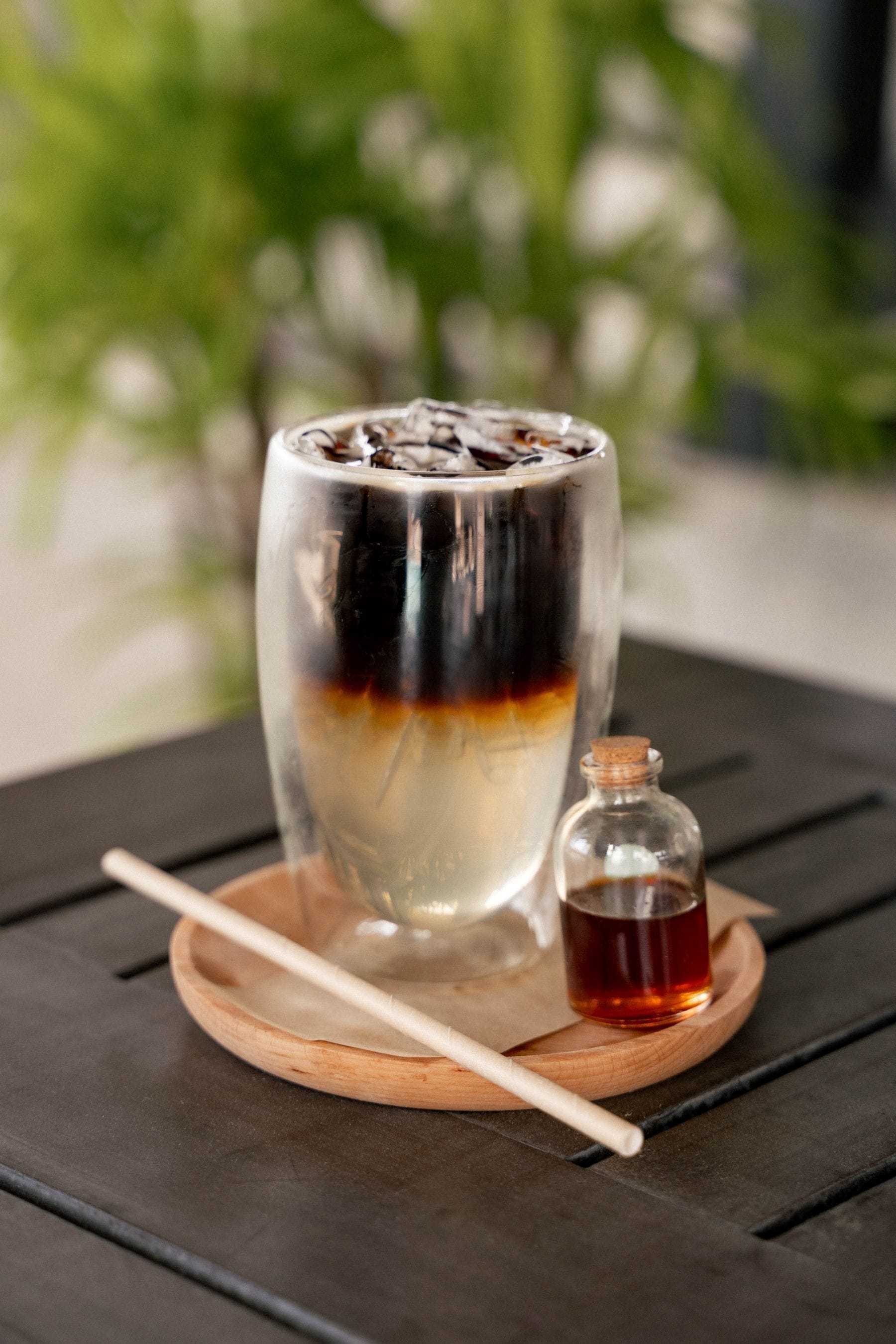 A tall glass with layered drinks on a wooden coaster, a small bottle of syrup, and a straw on a metal table.