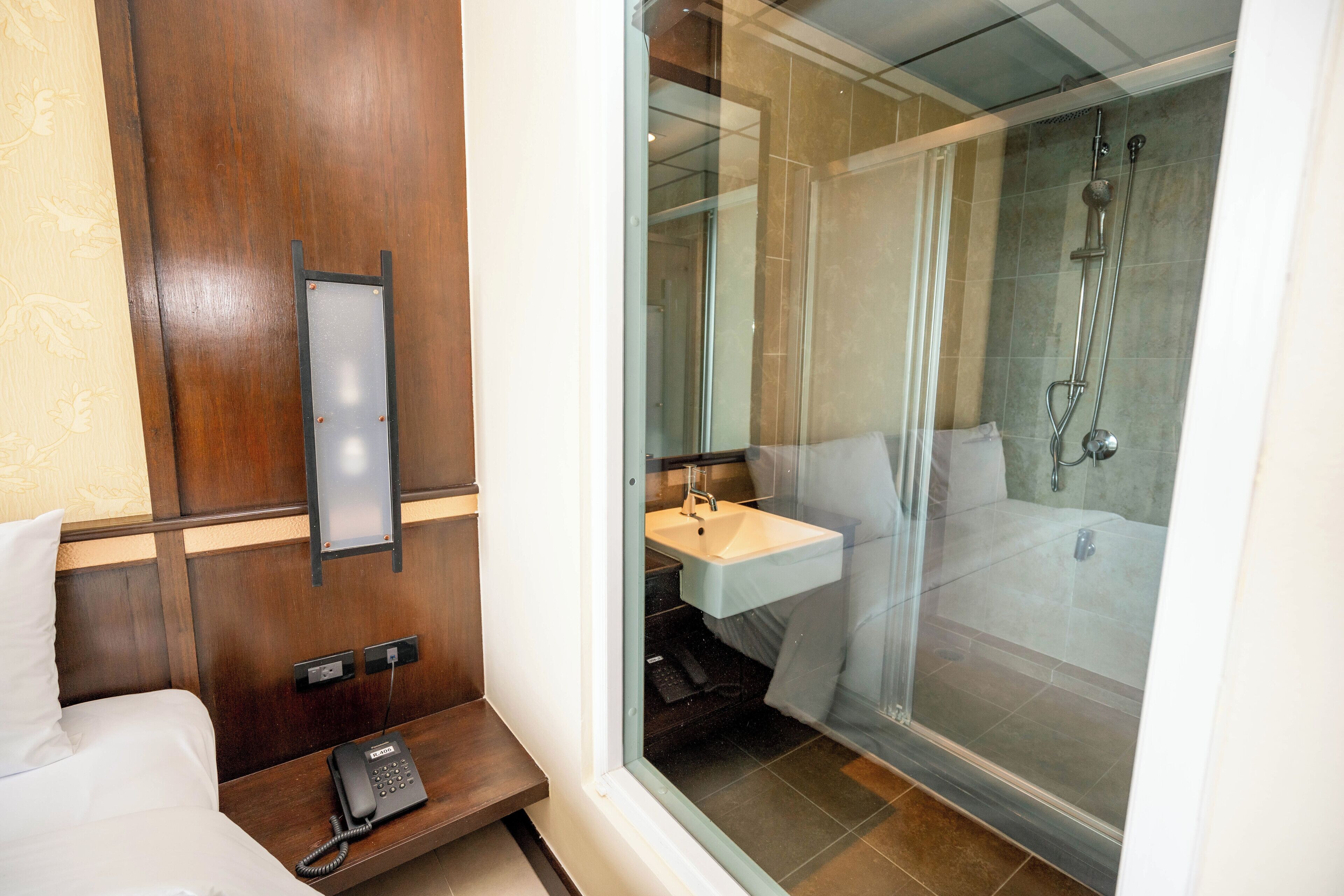 A hotel room with a wooden headboard, a bedside telephone, a glass-enclosed shower, and a sink.