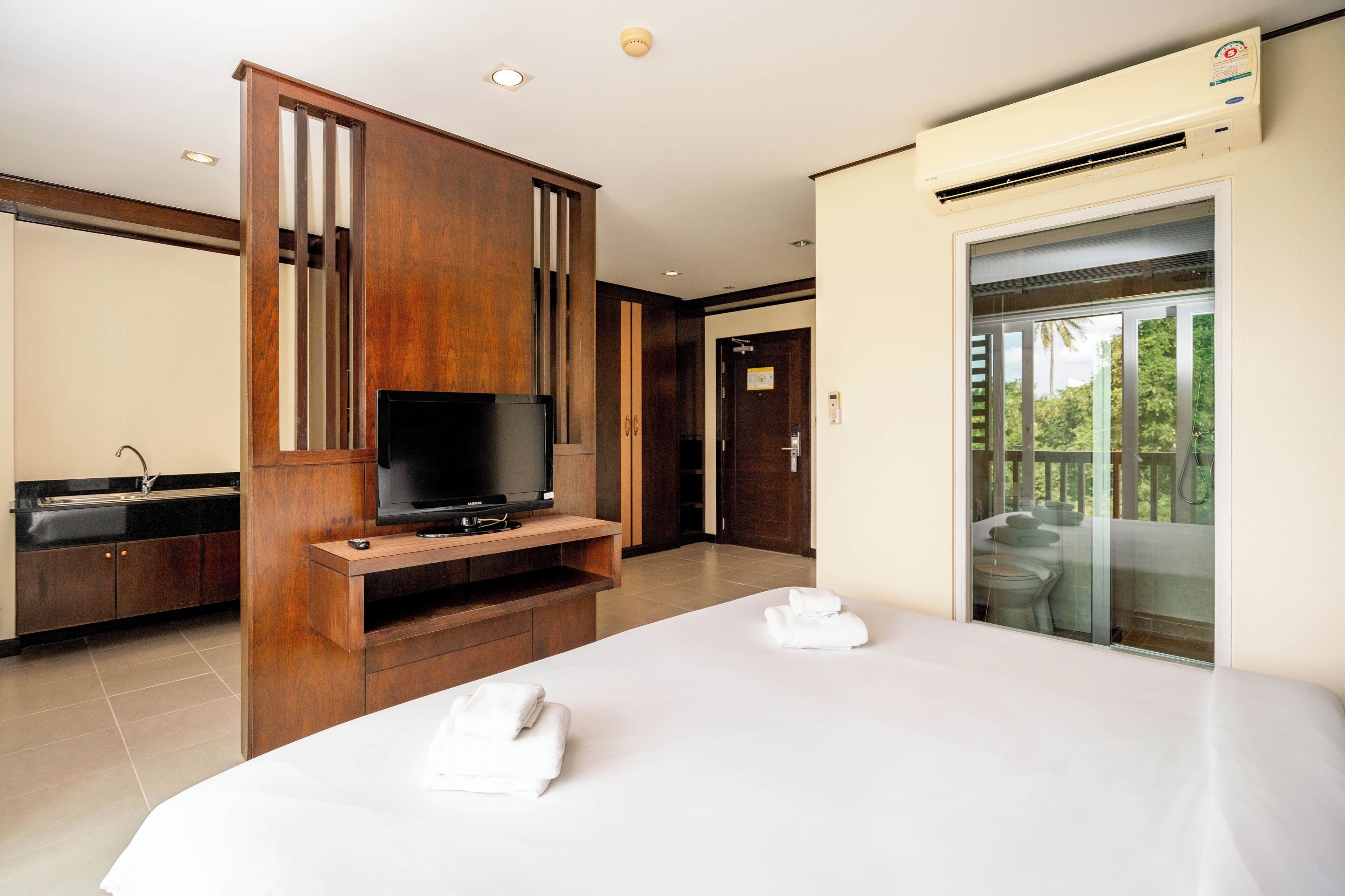 A hotel room with a large bed, a TV on a wooden stand, a balcony with a view of greenery, and a modern bathroom.