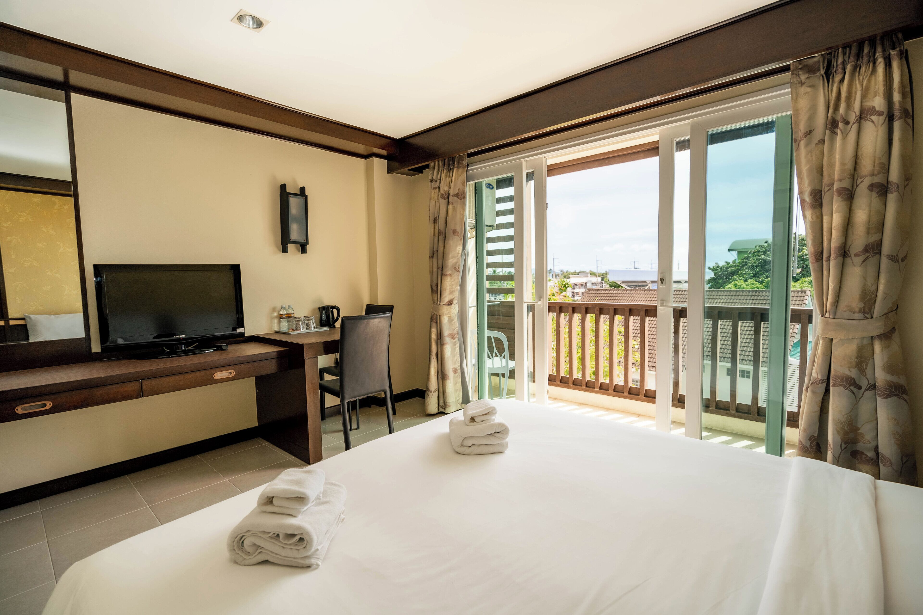 A hotel room with a bed, a desk with a TV, a chair, and a balcony with a view.