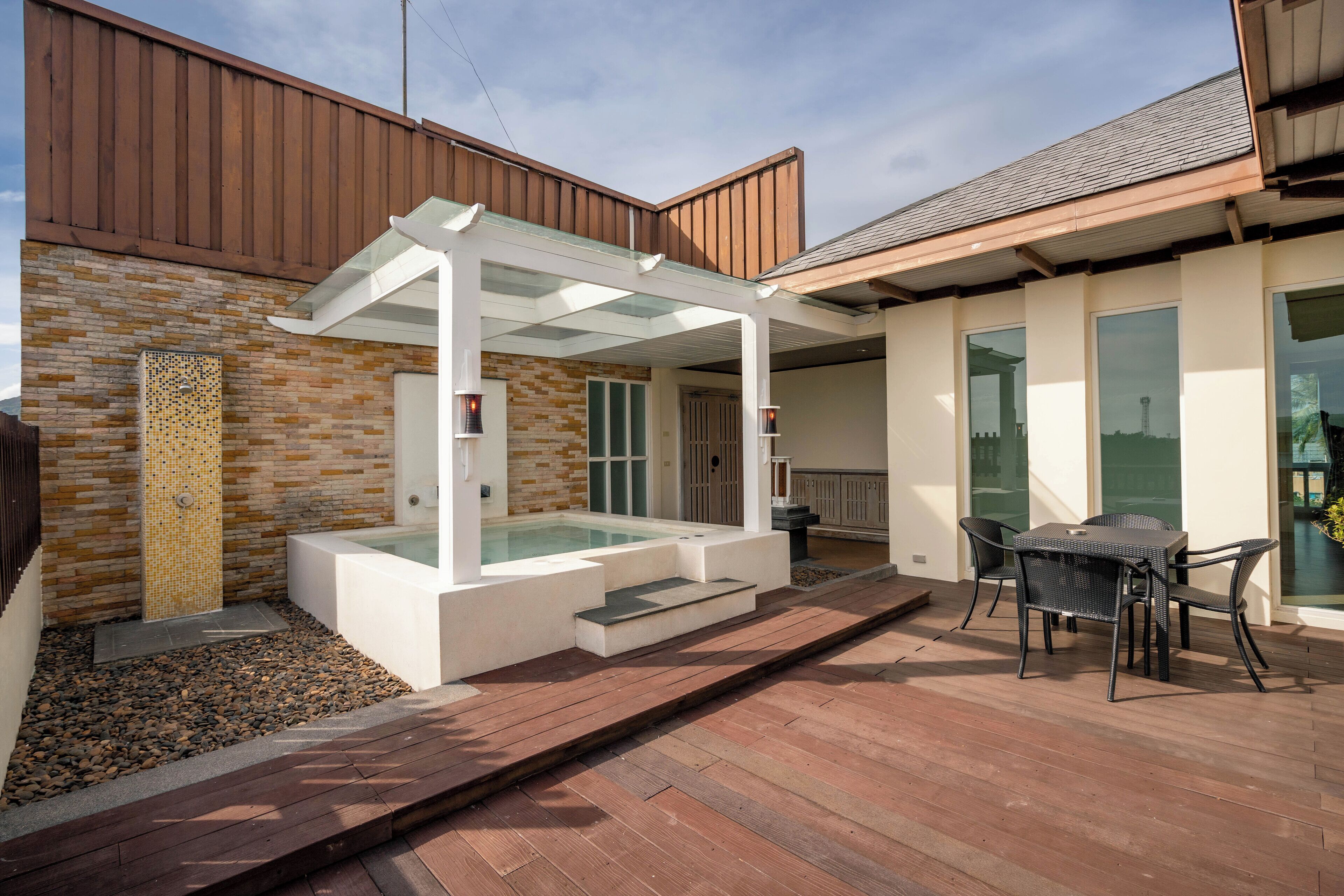 A modern outdoor patio with a glass-enclosed hot tub, wooden deck, and a dining area with chairs.