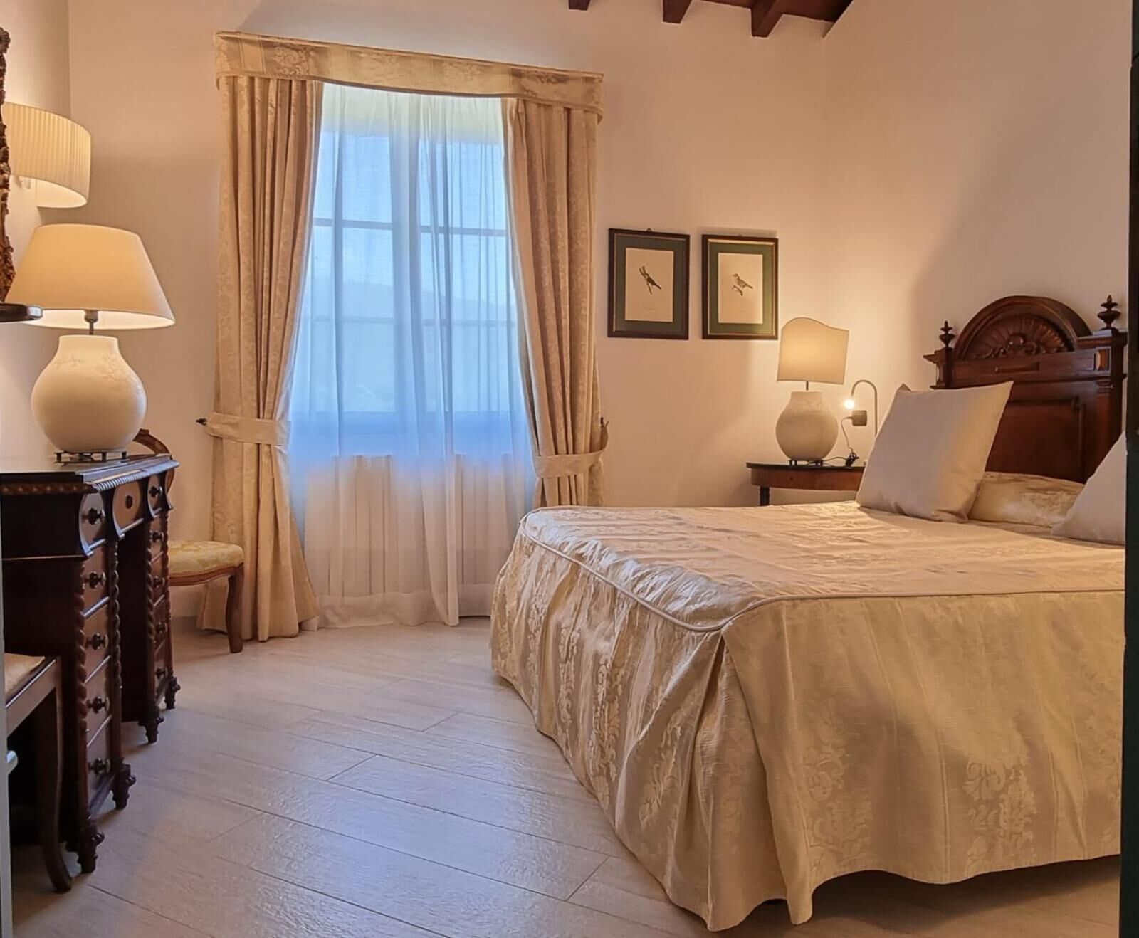 Un dormitorio con una cama grande, dos lámparas de noche, una mesita de madera, una silla y un ventanal con cortinas.