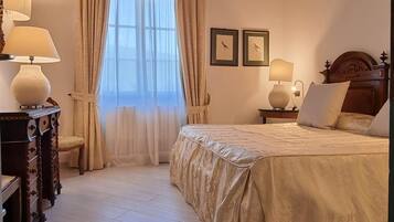 Un dormitorio con una cama grande, dos lámparas de noche, una mesita de madera, una silla y un ventanal con cortinas.