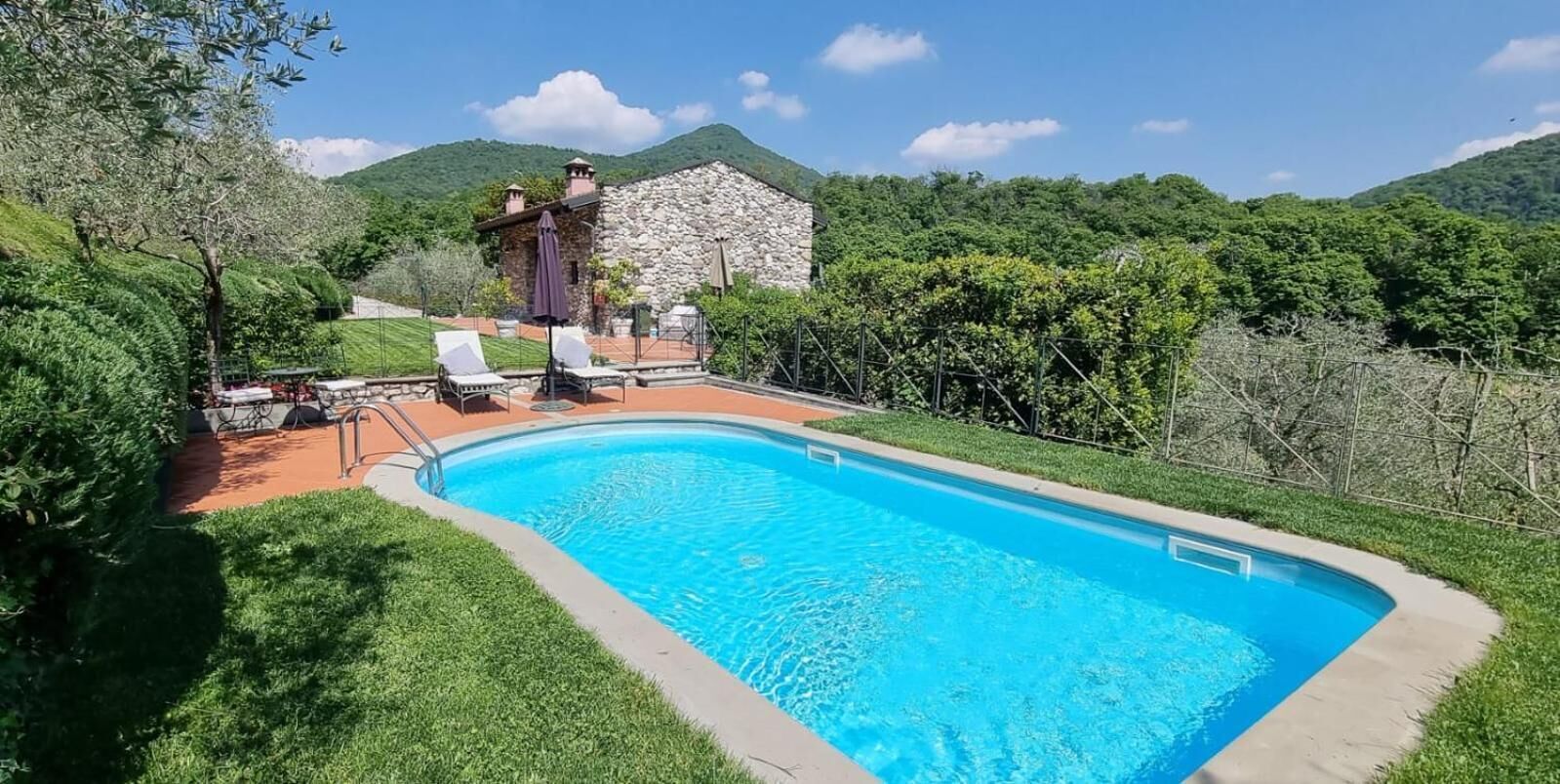 Una piscina con lettini, un edificio in pietra e vegetazione lussureggiante sullo sfondo.