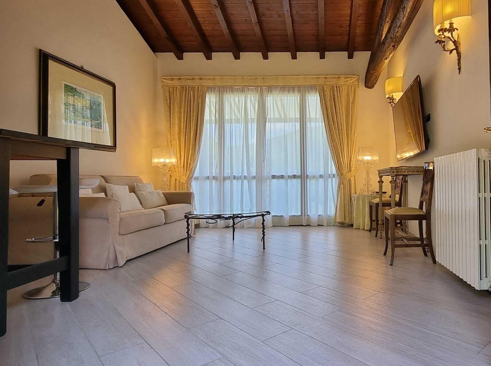 Een woonkamer met een bank, salontafel en een eettafel met stoelen. Er is een groot raam met gordijnen en een radiator aan de muur.