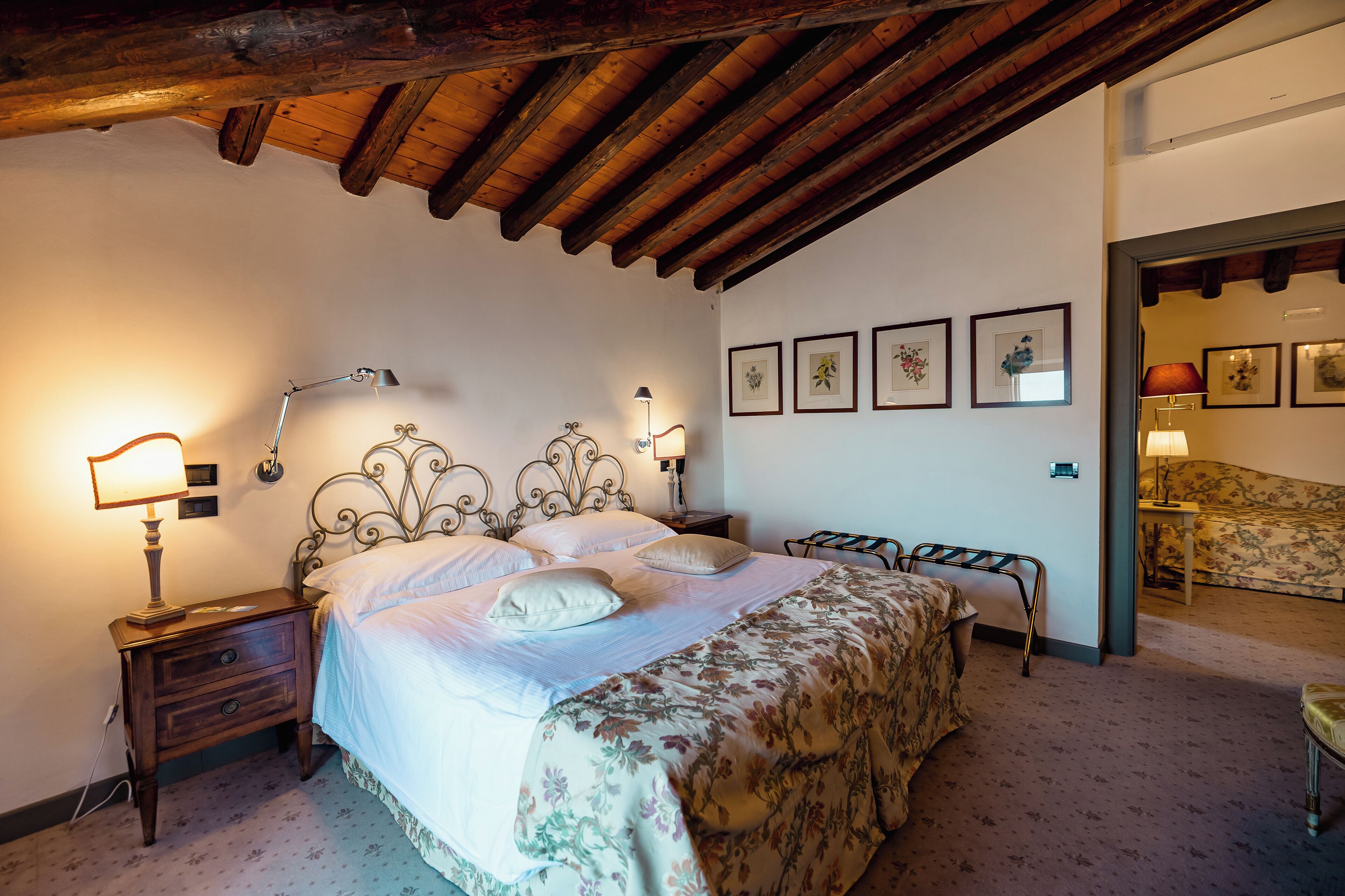 Un dormitorio con techo de madera, una cama con ropa de cama floral, lámparas de noche y cuadros en la pared.