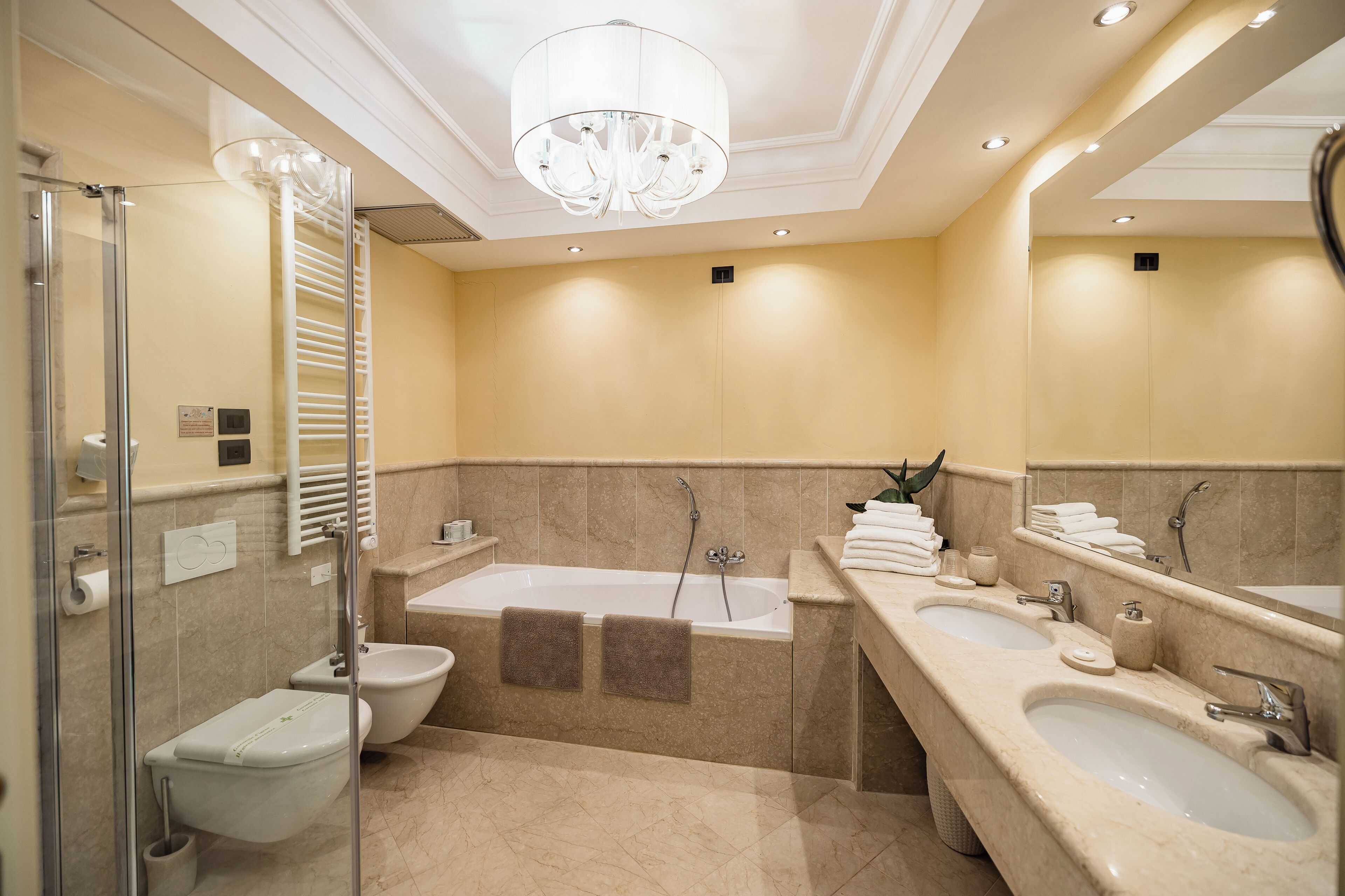 Une salle de bain moderne avec une douche à parois de verre, un bidet, une baignoire avec deux serviettes, un grand miroir et un lavabo double avec un meuble-vasque.
