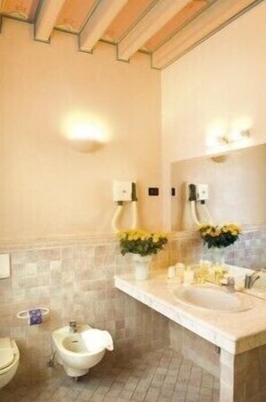 A bathroom with a toilet, bidet, and a sink with a mirror. There are two flower arrangements on the sink counter.