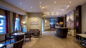 A hotel lobby with a reception desk, seating area, and a staircase leading to an upper floor.