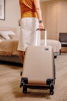 A person pulling a wheeled suitcase in a room with a bed and a chair.