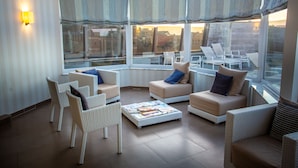 A sunlit room with wicker furniture, large windows, and a view of the city.