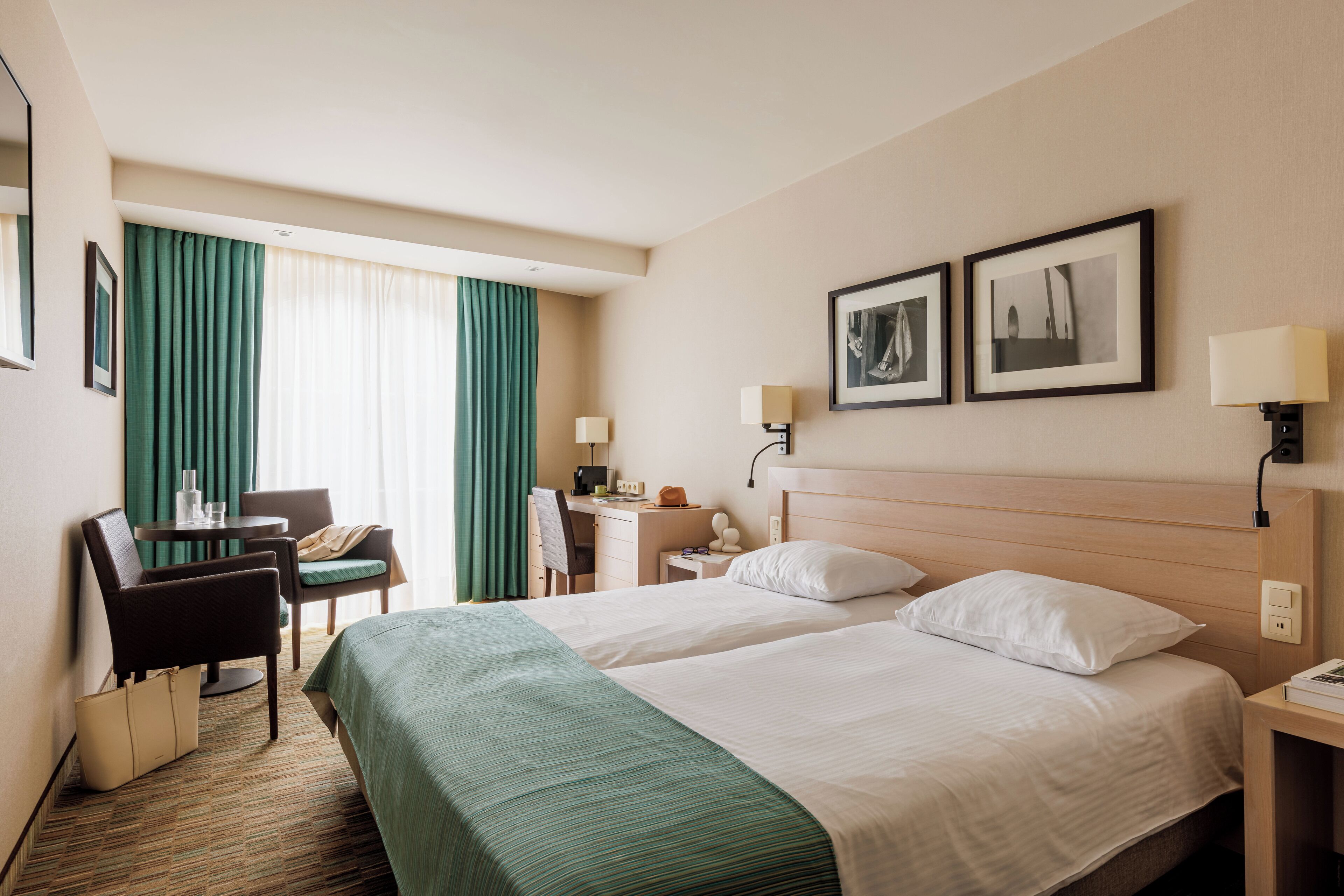 Une chambre d’hôtel avec deux lits, un bureau, une chaise, une lampe de bureau, un téléphone et deux tableaux encadrés au mur.