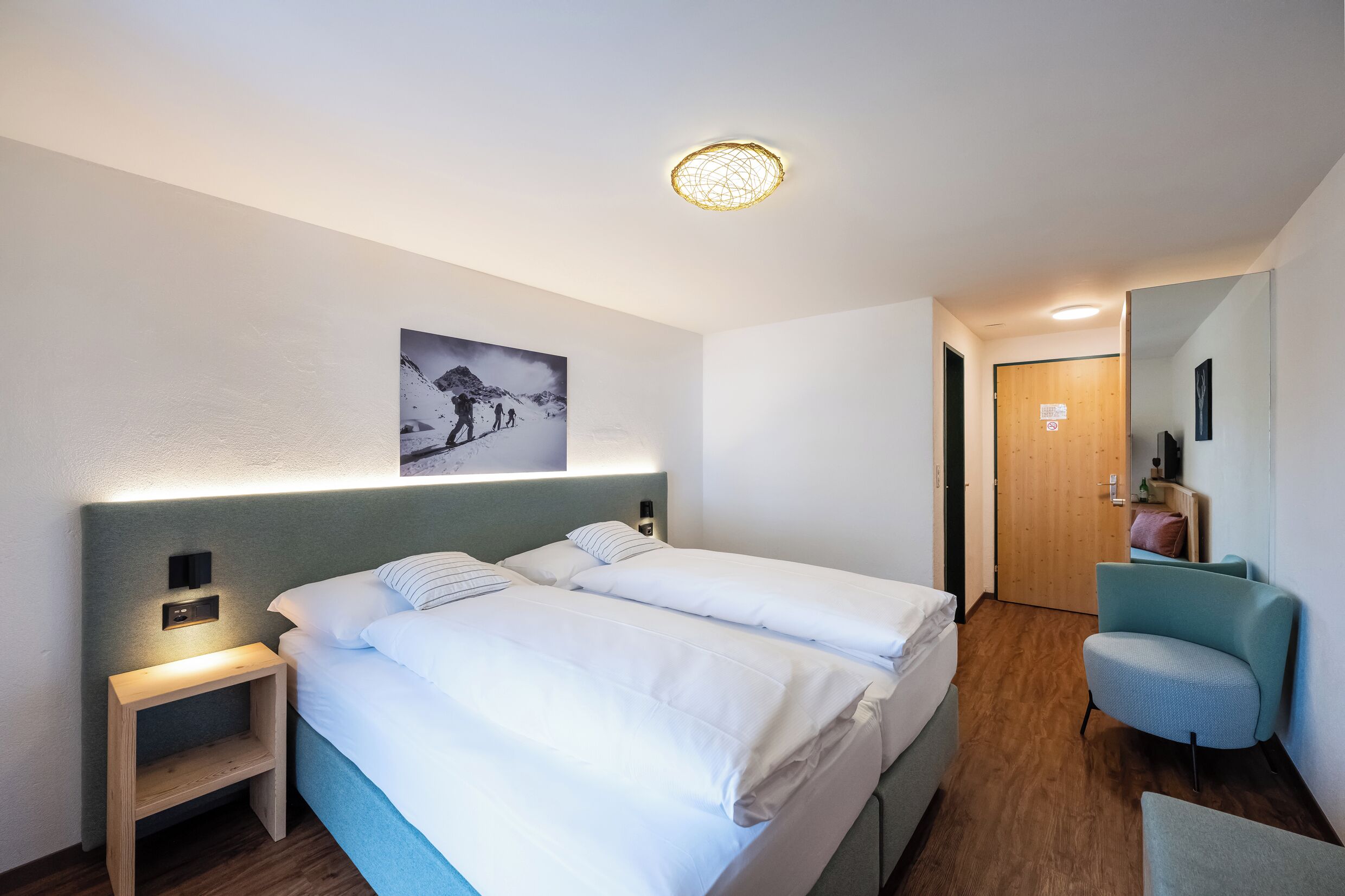 A hotel room with two beds, a nightstand, a chair, a wooden door, and a wall-mounted picture.