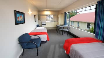 A hotel room with two single beds, a small kitchenette, a dining table, and a window with a view of a building.