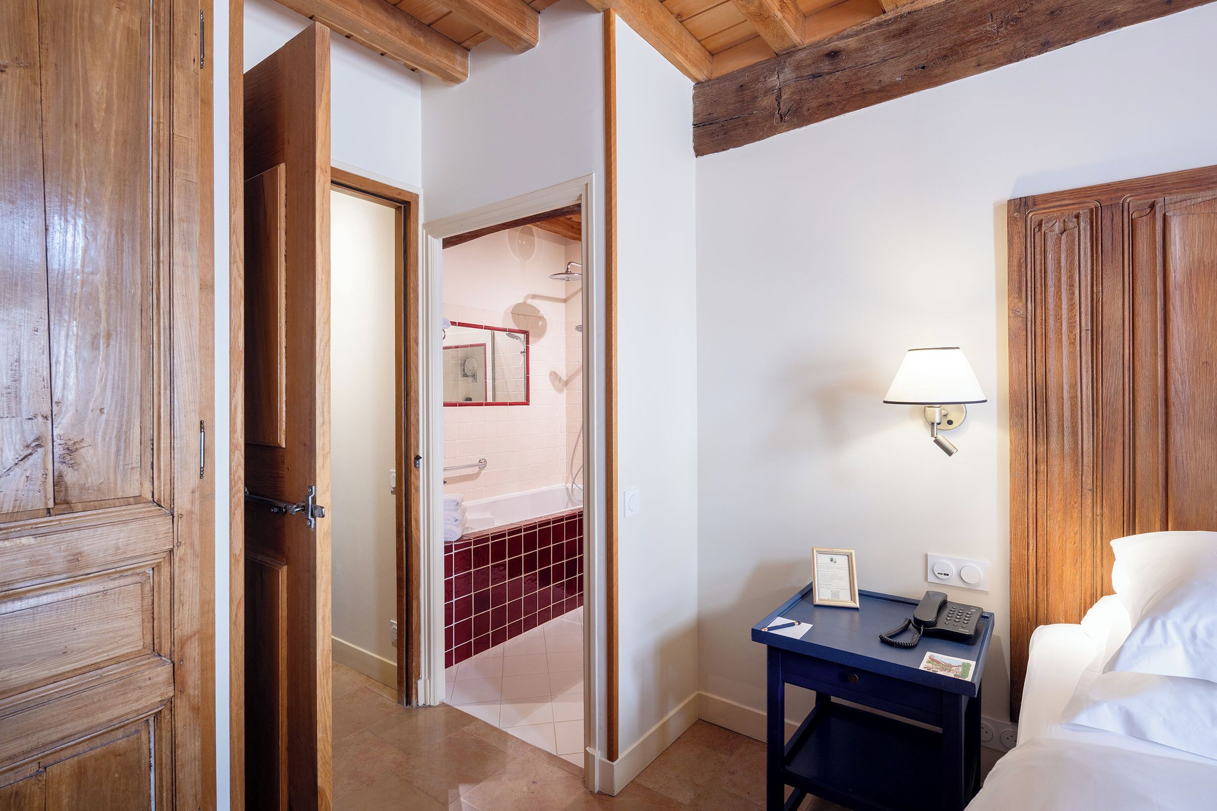A hotel room with a wooden door, a bathroom with a red-tiled shower, a bedside table with a lamp, and a white bed.