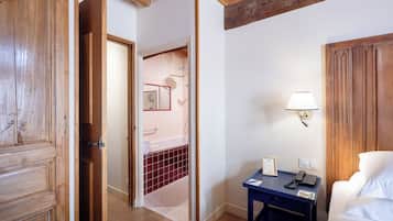 A hotel room with a wooden door, a bathroom with a red-tiled shower, a bedside table with a lamp, and a white bed.