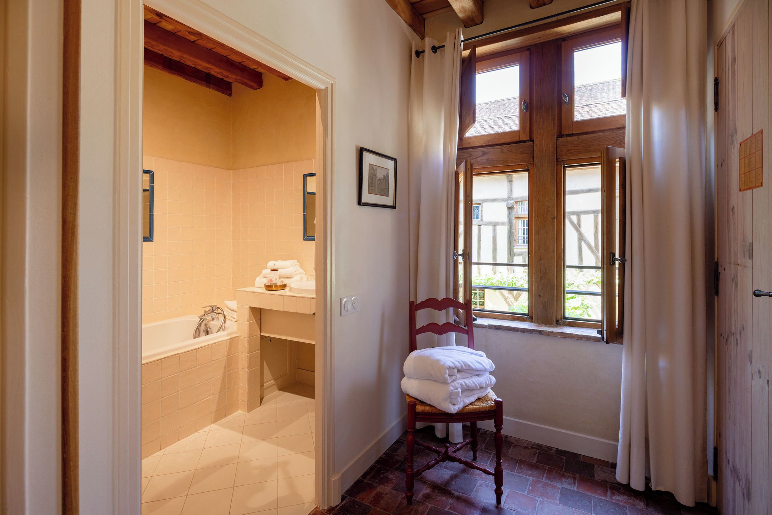 A bathroom with a bathtub, a chair with towels, and a window with curtains.