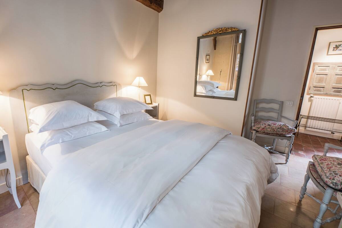 A neatly made bed with white linens, a bedside lamp, a chair with a floral cushion, and a mirror on the wall.