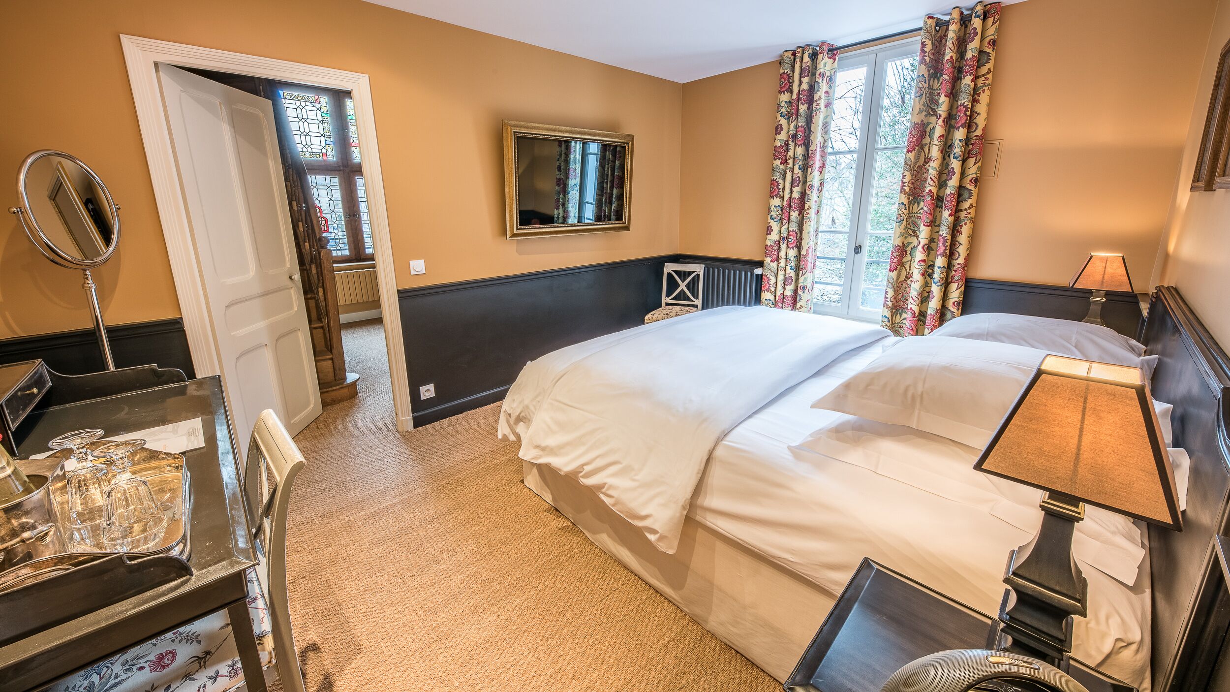 Een hotelkamer met een bed, een bureau met spiegel, een dressoir, een raam met bloemen gordijnen en een kleine tafel met lamp.