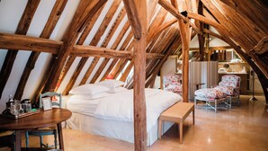 A cozy attic bedroom with a wooden ceiling, a bed with white linens, a wooden bench, a chair with floral upholstery, and a small table with a tray.