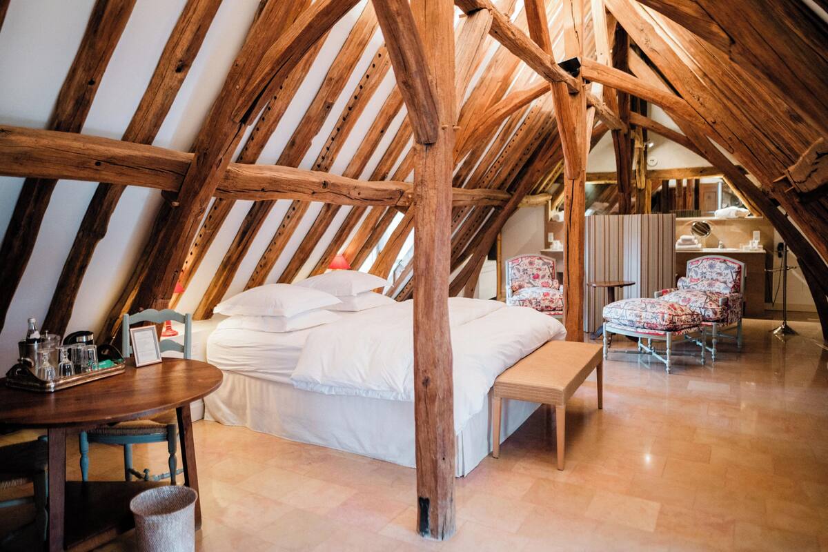 A cozy attic bedroom with a wooden ceiling, a bed with white linens, a wooden bench, a chair with floral upholstery, and a small table with a tray.