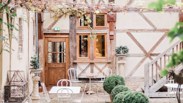 A traditional half-timbered building with a wooden door, a small balcony, and a stone staircase. There are white metal chairs and a table in front of the building.