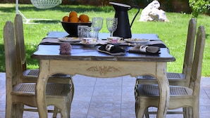 An outdoor dining area with a table set for four, surrounded by chairs, a hanging light fixture, and a view of a lush garden and distant hills. - SunVillage Boutique Hotel (Hersonissos)