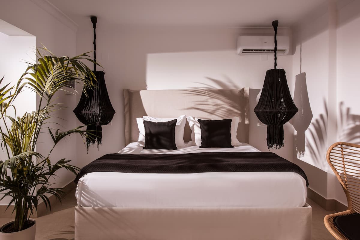 Ein modernes Schlafzimmer mit einem großen Bett, zwei hängenden schwarzen Lampen, einem Blumentopf und einem Korbstuhl.