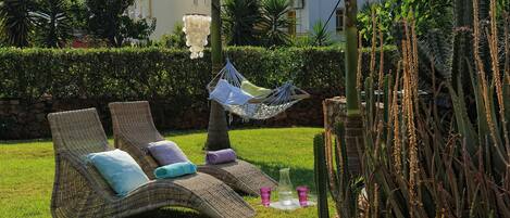 Two wicker lounge chairs with cushions, a hanging hammock, and a cactus plant in a grassy outdoor area.