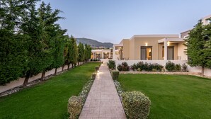 A modern hotel with a paved pathway leading to the entrance, surrounded by well-maintained greenery and a mountain backdrop. - Plakias Suites (Agios Vasileios)