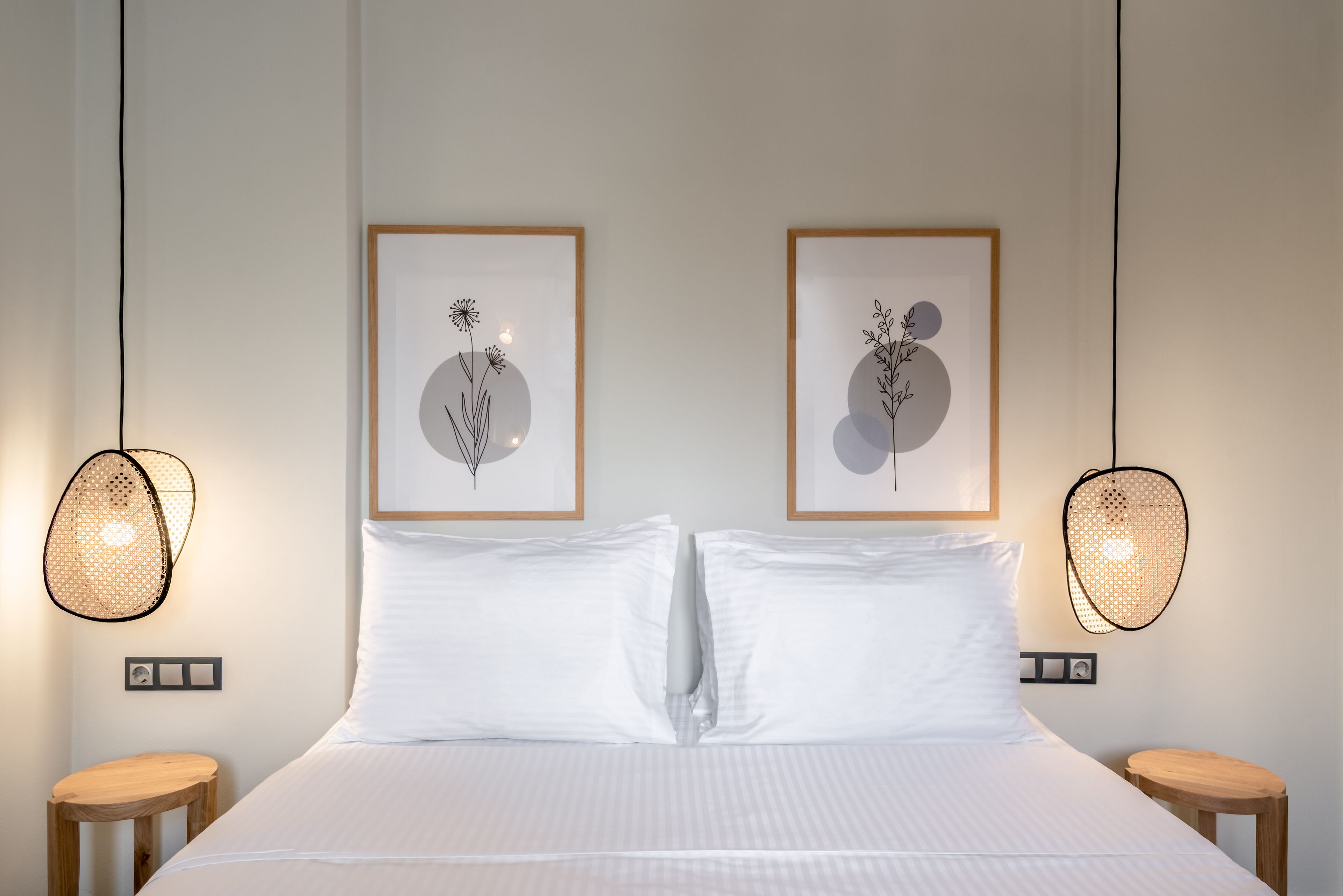 Een net opgemaakt bed met witte lakens, twee ingelijste kunstwerken met bloemen en twee rieten lampen aan weerszijden.
