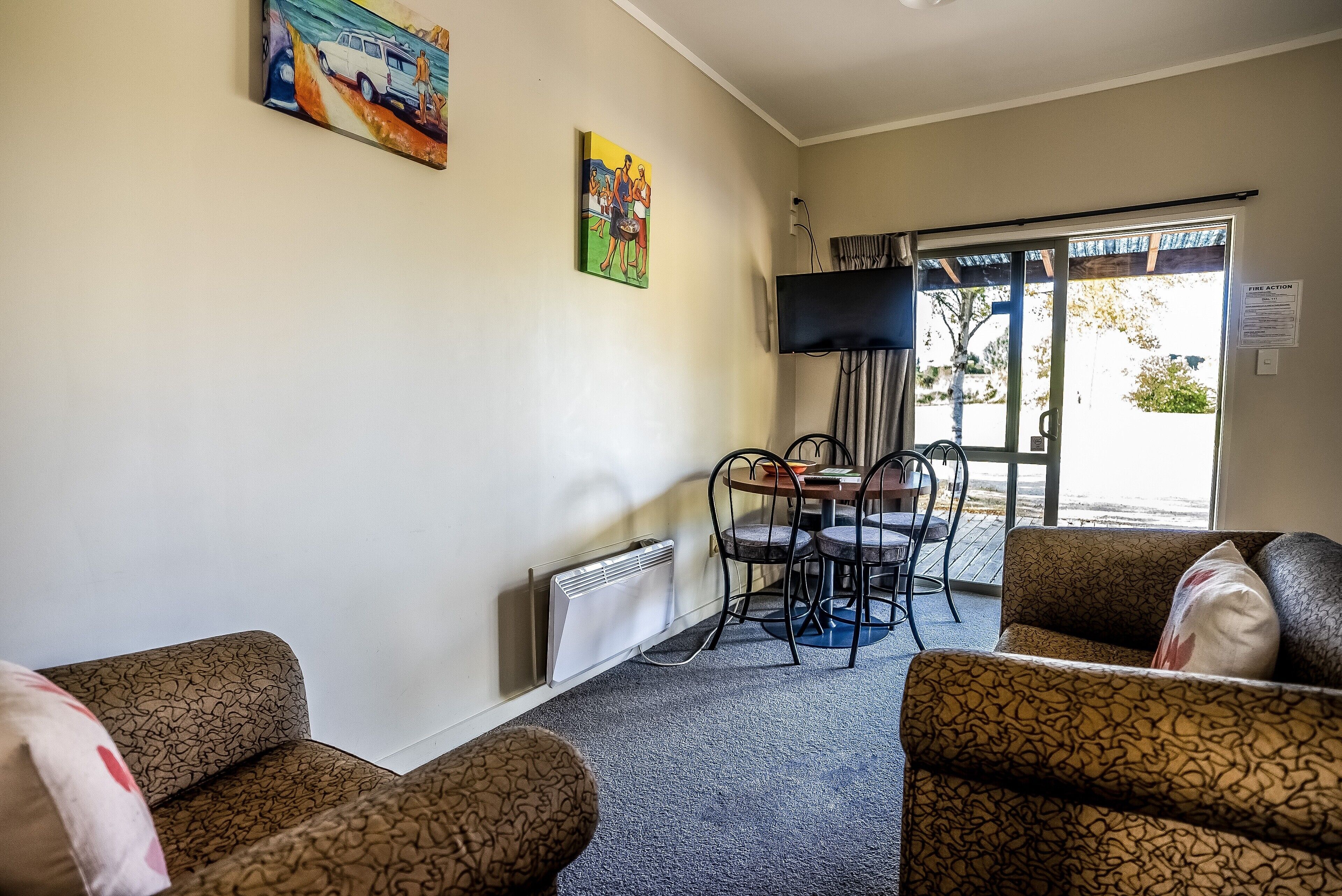 A room with patterned sofas, a table with chairs, a TV mounted on the wall, and a sliding glass door leading outside.