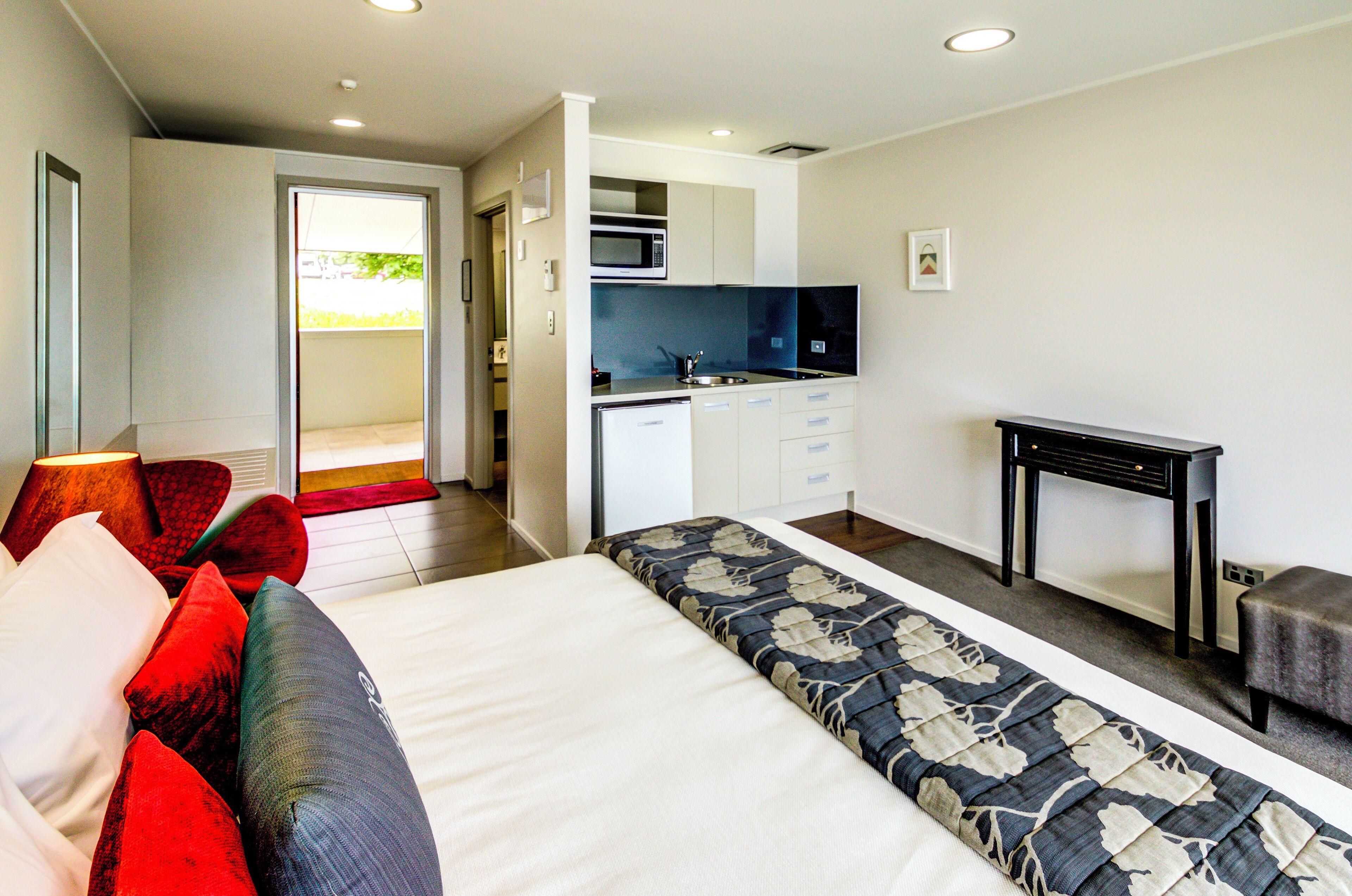 A modern hotel room with a large bed, a desk, a kitchenette, and a view of the outdoors.
