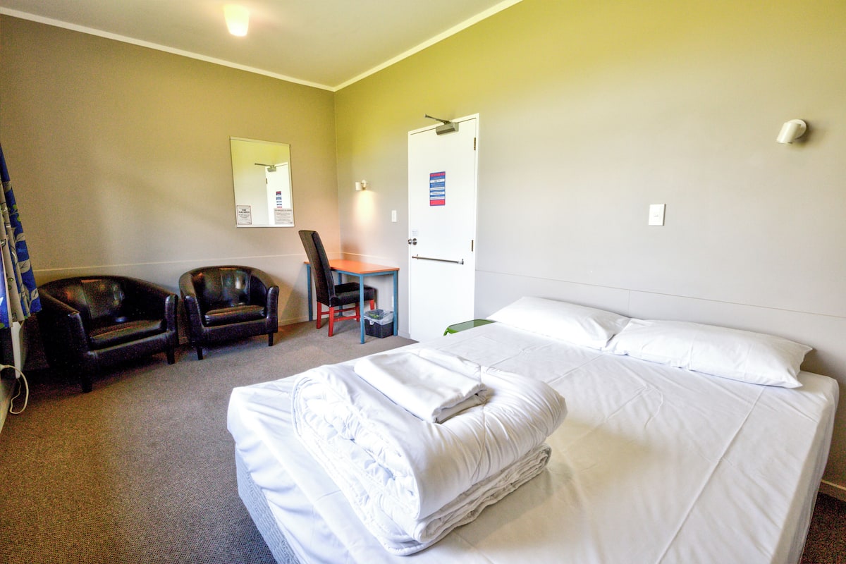 A hotel room with a bed, two chairs, a desk, and a door.