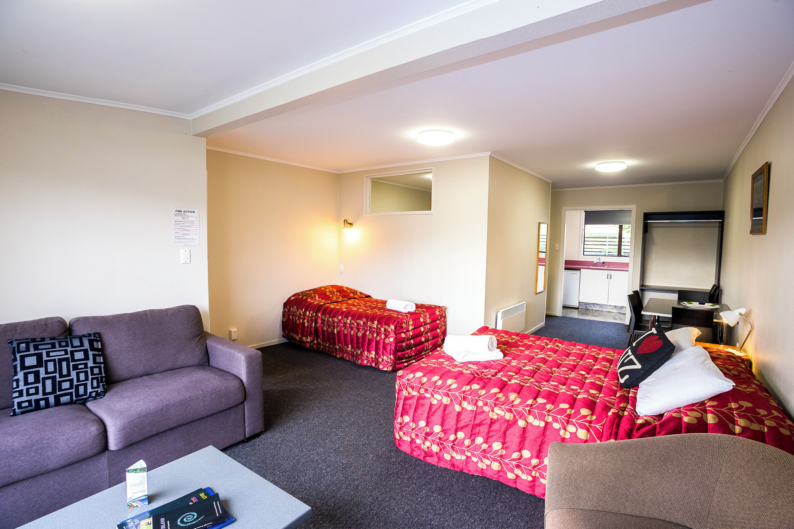 Habitación de hotel con sofá, dos camas, una mesita y una kitchenette al fondo.