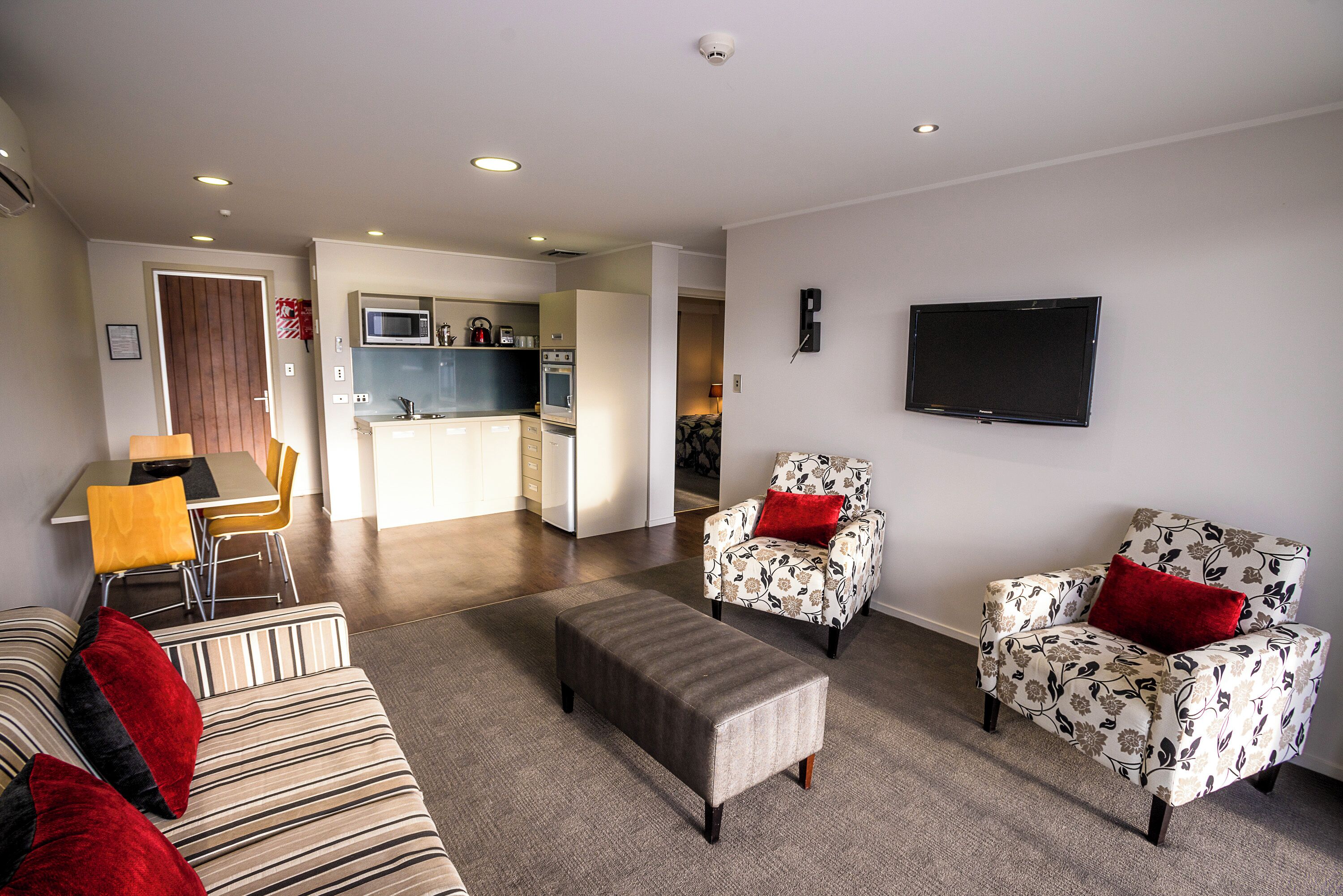 A modern living room with a sofa, ottoman, and floral armchairs, a flat-screen TV, and a kitchen area in the background.