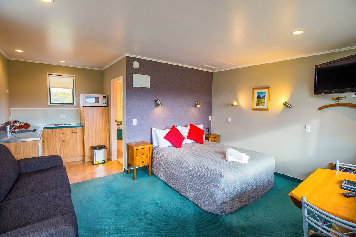 A hotel room with a bed, a sofa, a desk, a TV, a microwave, and a small kitchen area.