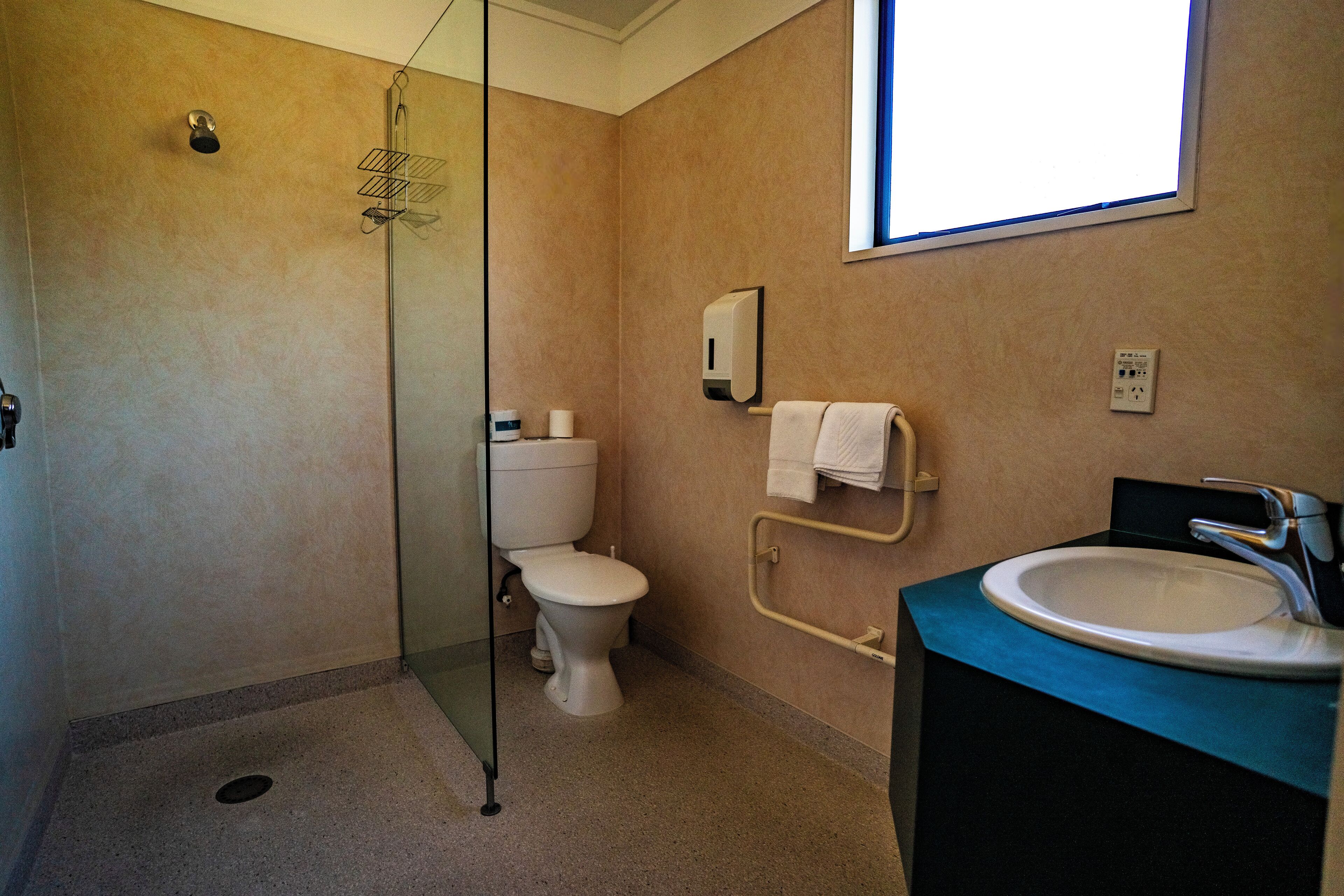 Ein Badezimmer mit Duschkabine aus Glas, Toilette, Waschbecken und Handtuchhalter sowie einem Fenster für Tageslicht.