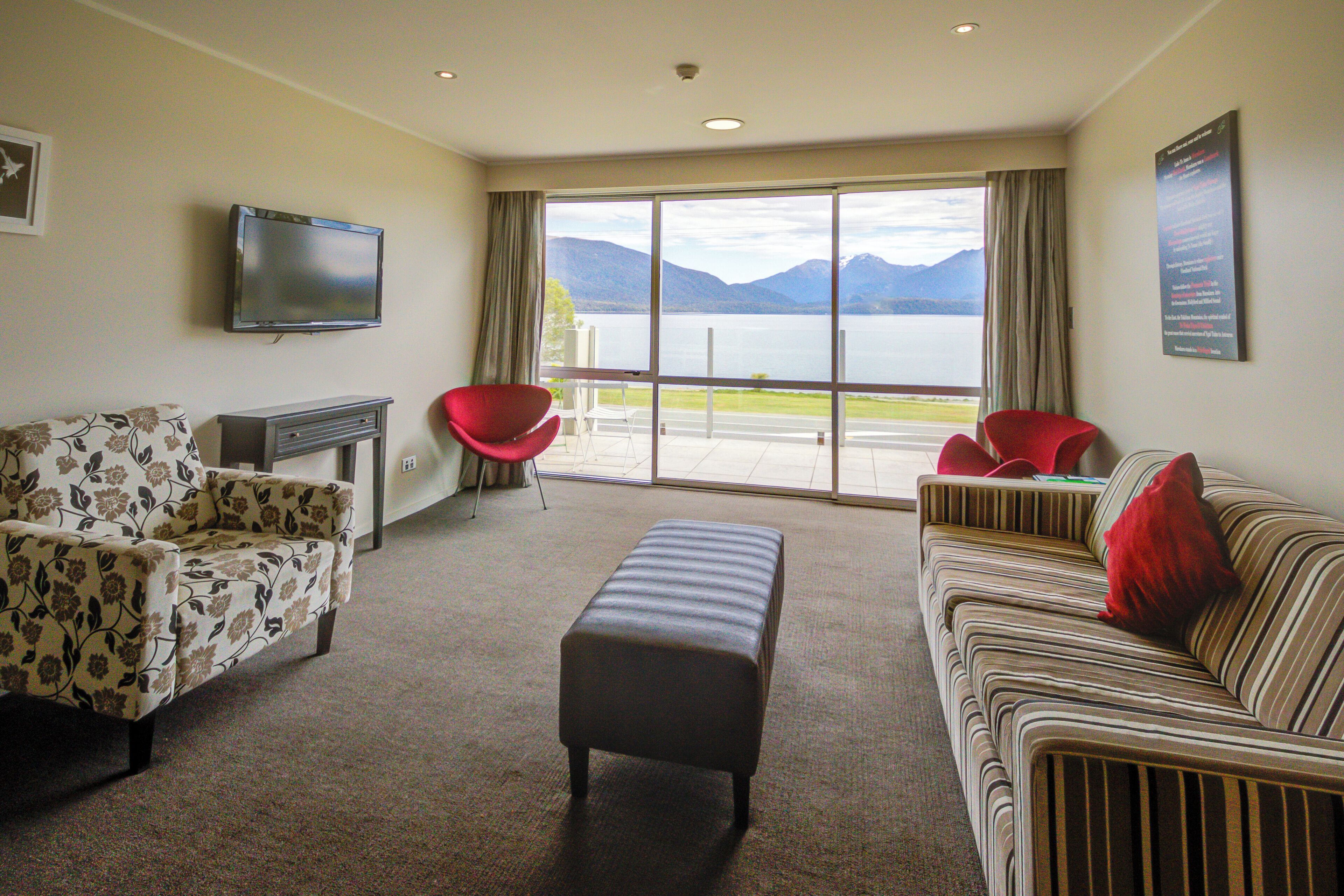 A living room with a sofa, ottoman, and chairs, a flat-screen TV, and a large window with a view of mountains and a lake.