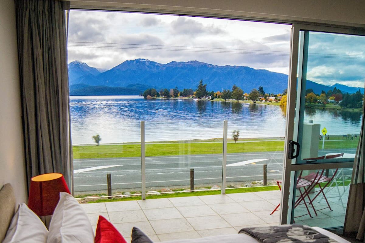 A bedroom with a large bed, a view of a lake and mountains, and a balcony with a chair.