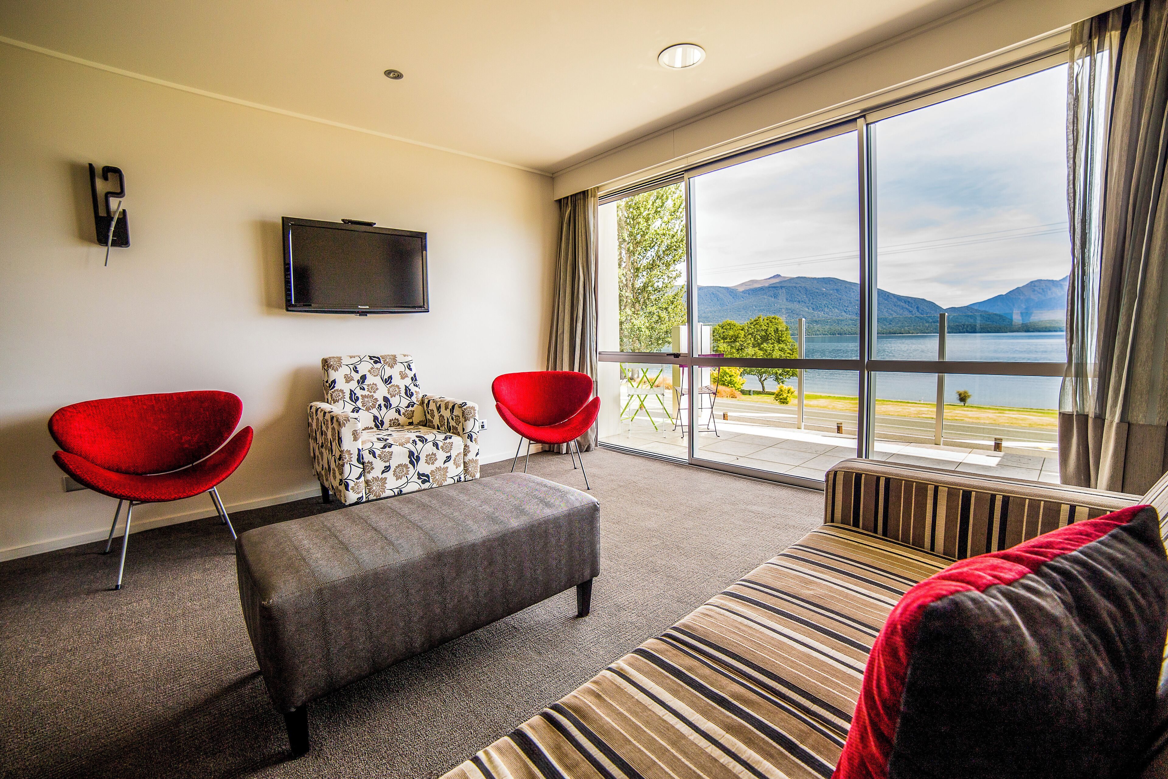 Een moderne hotelkamer met een groot raam dat uitzicht biedt op een meer en bergen. Er is een flatscreen televisie aan de muur, een grijze bank, een rode stoel en een fauteuil met een patroon.