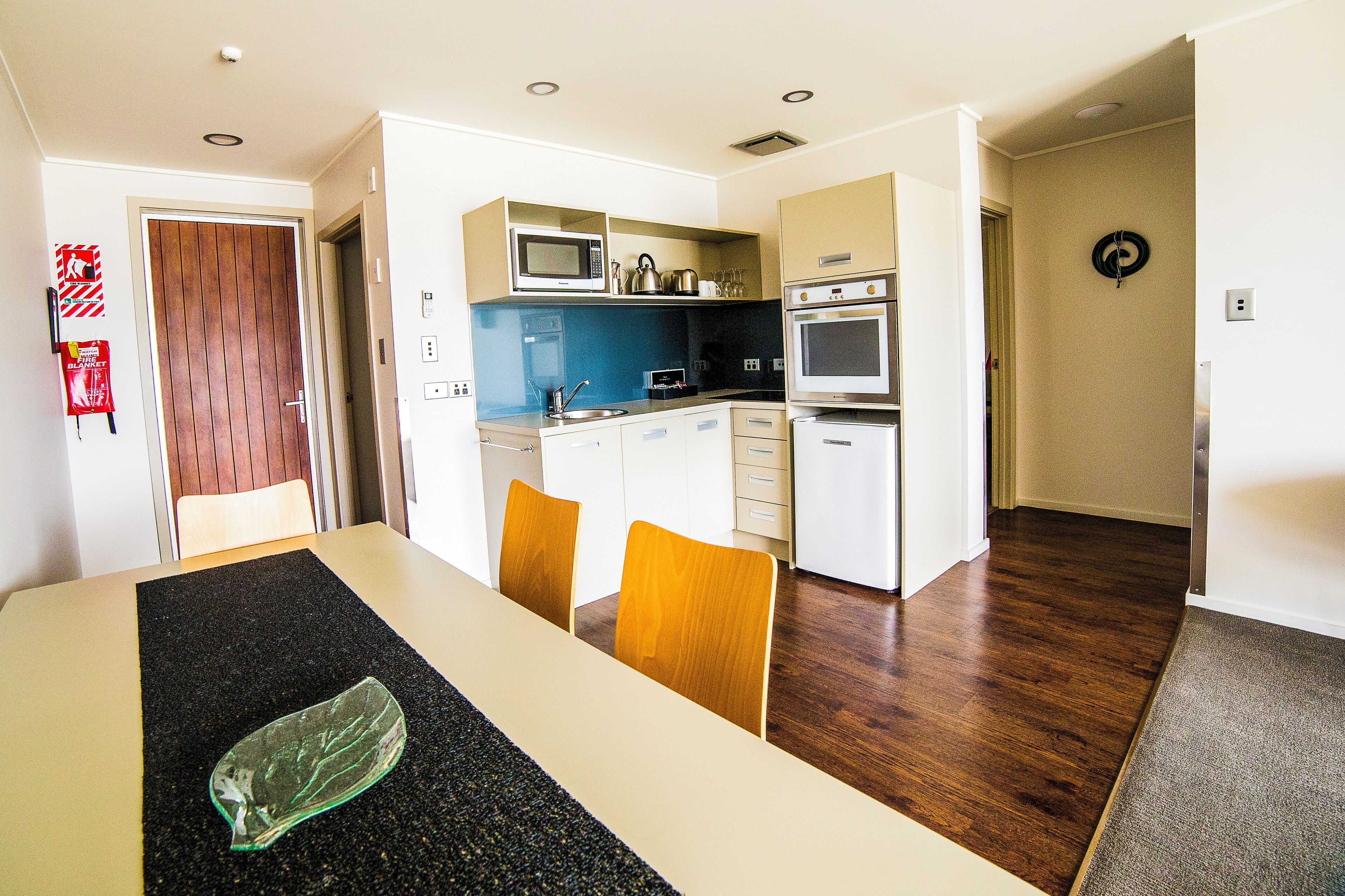 A modern kitchen with a microwave, oven, and kettle. There is a dining table with chairs and a black placemat.