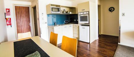 A modern kitchen with a microwave, oven, and kettle. There is a dining table with chairs and a black placemat.