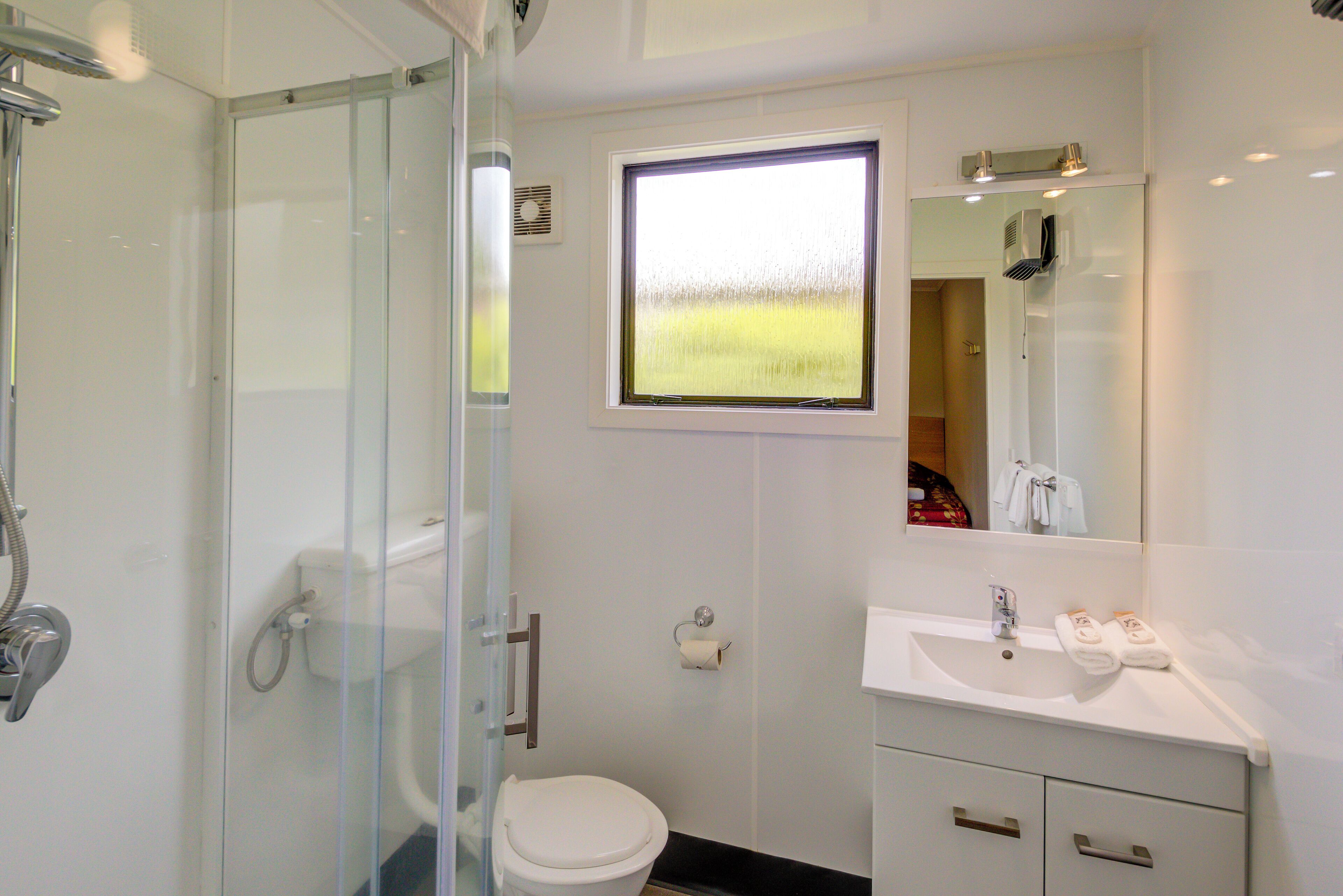 A modern bathroom with a glass shower enclosure, a toilet, a sink with a mirror, and a window with a view of a field.
