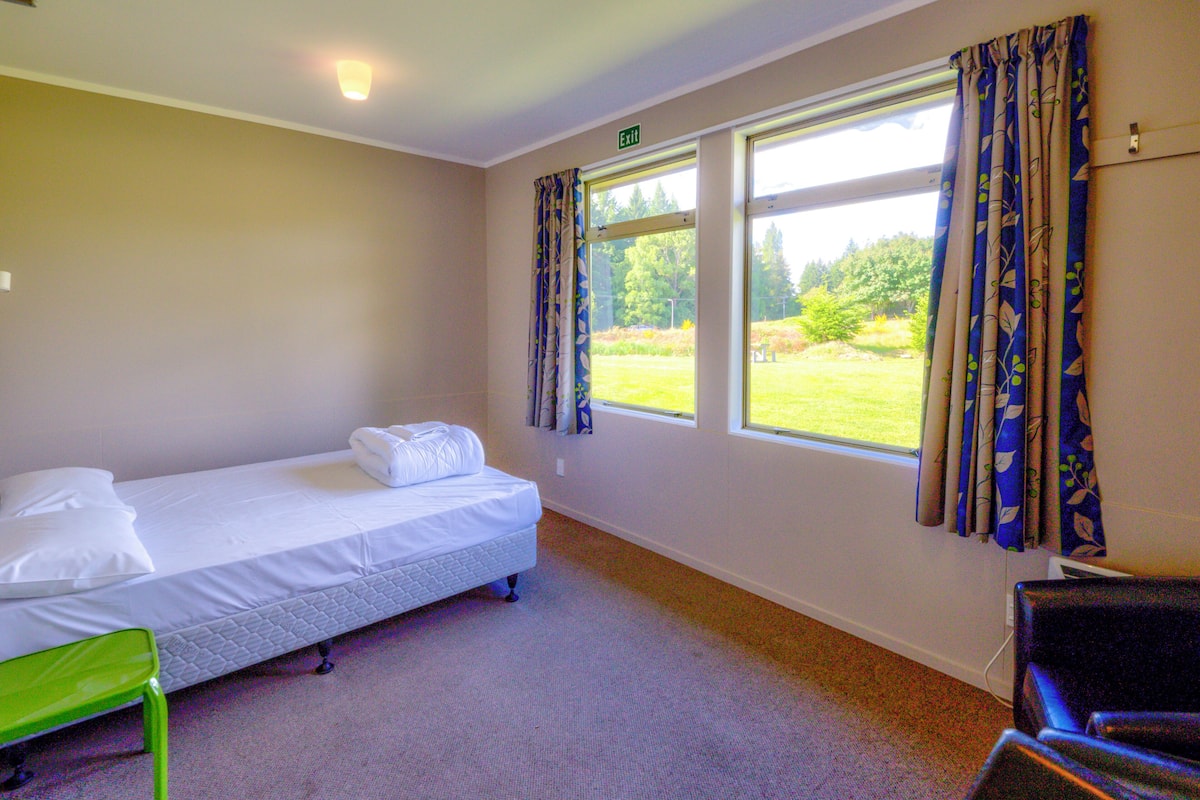 A single-bed room with a green chair, a window with curtains, and an exit sign.