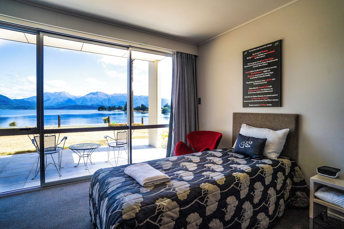 A hotel room with a large bed, a red chair, a small table, and a view of a lake and mountains.